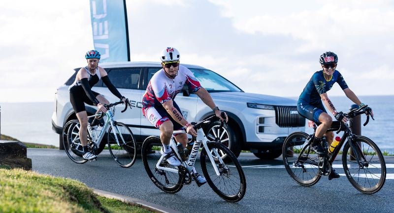 2025 IRONMAN competitors cycle alongside JAECOO vehicle