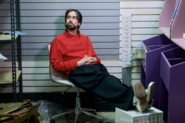 Alex G sitting in an office chair looking to the left, in a messy office space