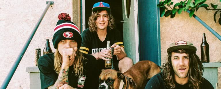Three members of band Dune Rats sitting outside a house on the steps, drinking beer, with a boxer dog, wearing caps and a San Francisco beanie