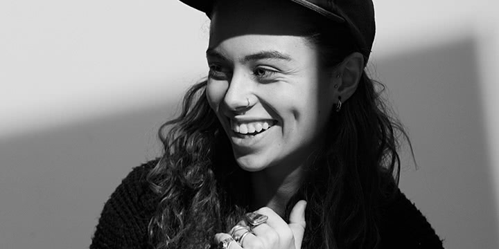 black and white portrait of Tash Sultana smiling, wearing a black cap and three rings on her fingers and a nosering