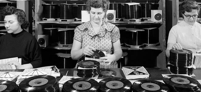 Black and white vintage photo of three women working in a vinyl pressing factory, packaging 7" singles into record sleeves