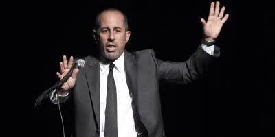 Stand-up comedian Jerry Seinfeld on-stage telling a joke, gesticulating wildly whilst holding a microphone