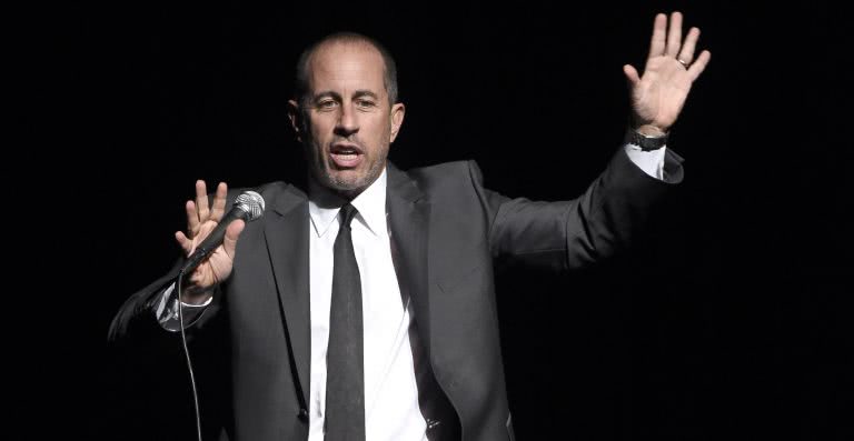 Stand-up comedian Jerry Seinfeld on-stage telling a joke, gesticulating wildly whilst holding a microphone