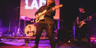 Alex Lahey performing at Electric Lady, mouth open as she shreds on guitar in front of a screen bearing the event's title.