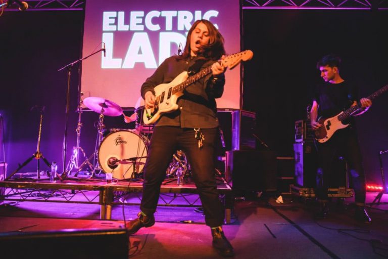 Alex Lahey performing at Electric Lady, mouth open as she shreds on guitar in front of a screen bearing the event's title.