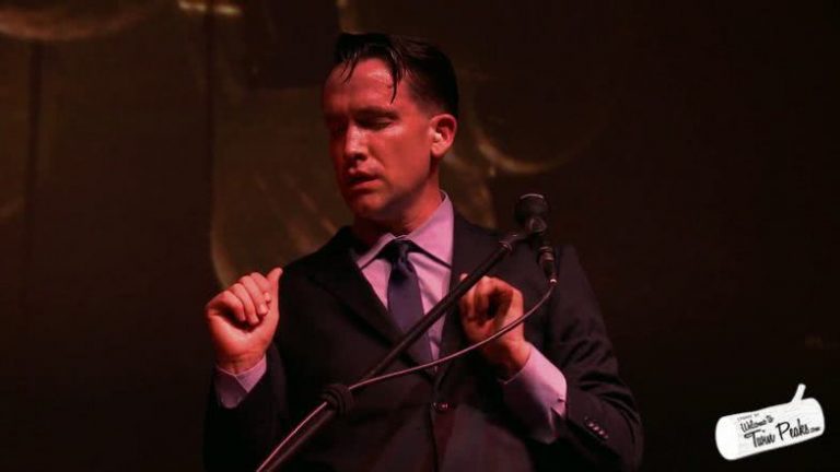 Jamie Stewart of Xiu Xiu, wearing a suit and standing with eyes closed behind a keyboard and microphone.