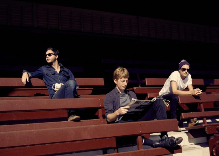 The three members of Fountaineer, one reading a newspaper, sitting in the stands of a suburban sportsfield