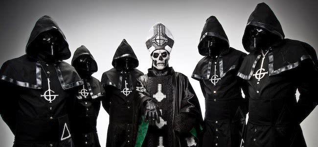 5 men standing in hooded costumes with faces covered and man standing in the middle with a skull mask in a black cloak