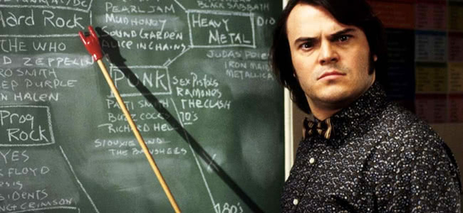 Jack Black looking at the camera in a black and white shirt standing in front of a black board