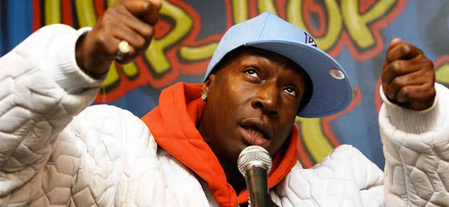 Man wearing a black and red jumper and a blue hat talking into a microphone with his hands in the air