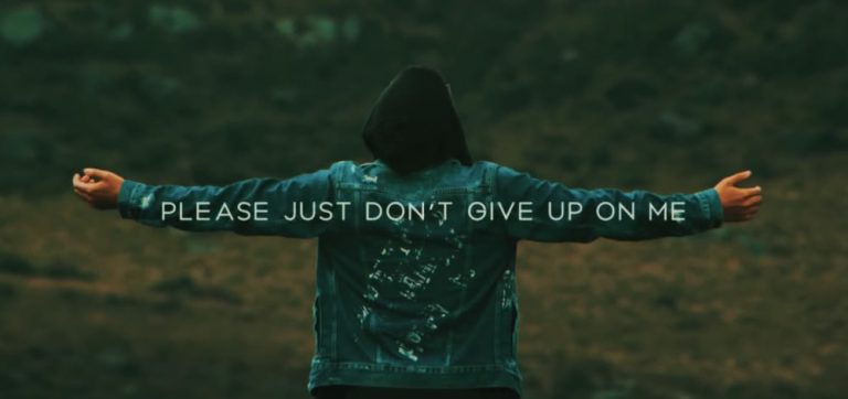 Man standing in a green field wearing a denim hoodie with his back to the camera and arms stretched out with writing across his arm span