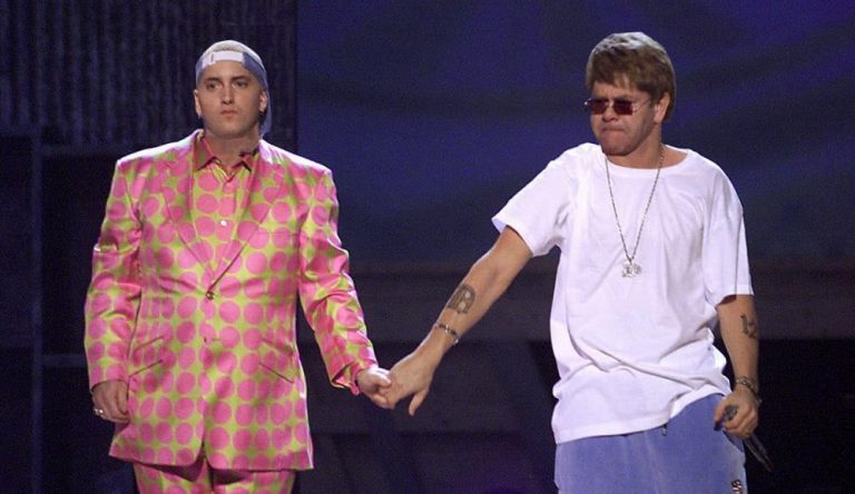 Eminem wearing a pink and green spotted suit holding hands with Elton John who is wearing a white tshit and blue tracksuit pants