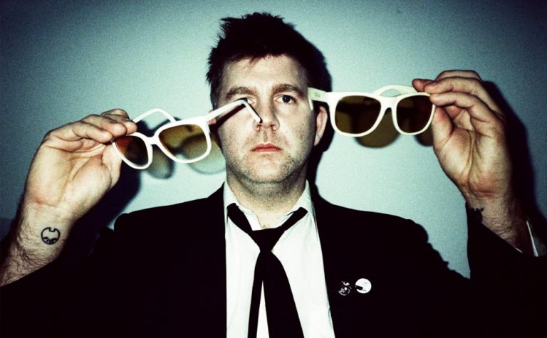 Man in black suit, whist shit and black tie holding two pairs of white sunglasses with a tattoo on his write looking at the camera