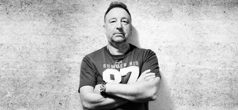 Black and white image of Peter Hook standing against a wall with his arms crossed
