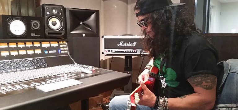 Slash wearing a backwards cap, playing a guitar sitting in a recording studio