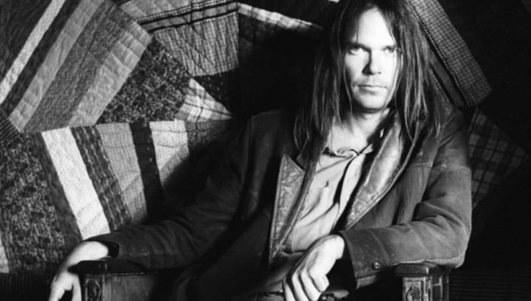 Neil Young sitting on a chair with a dark jacket on looking at the camera