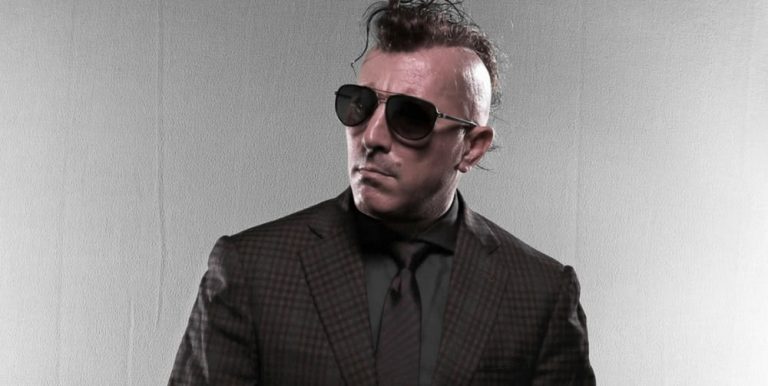Maynard James Keenan standing in a black suit wearing dark sunglasses